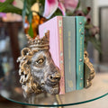 Three decorative books standing upright in a gold-toned lion head bookend on a glass table, with a blurred indoor background and flowers behind.
