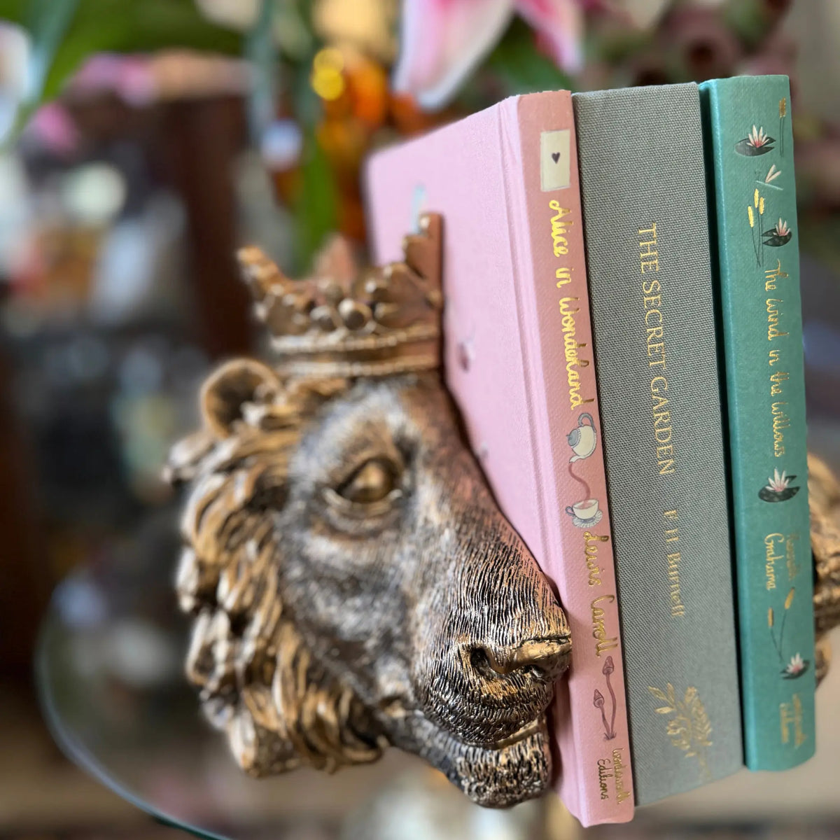A decorative bronze lion head bookend holding three books on a reflective glass table. The books have pink, gray, and teal spines with gold lettering.