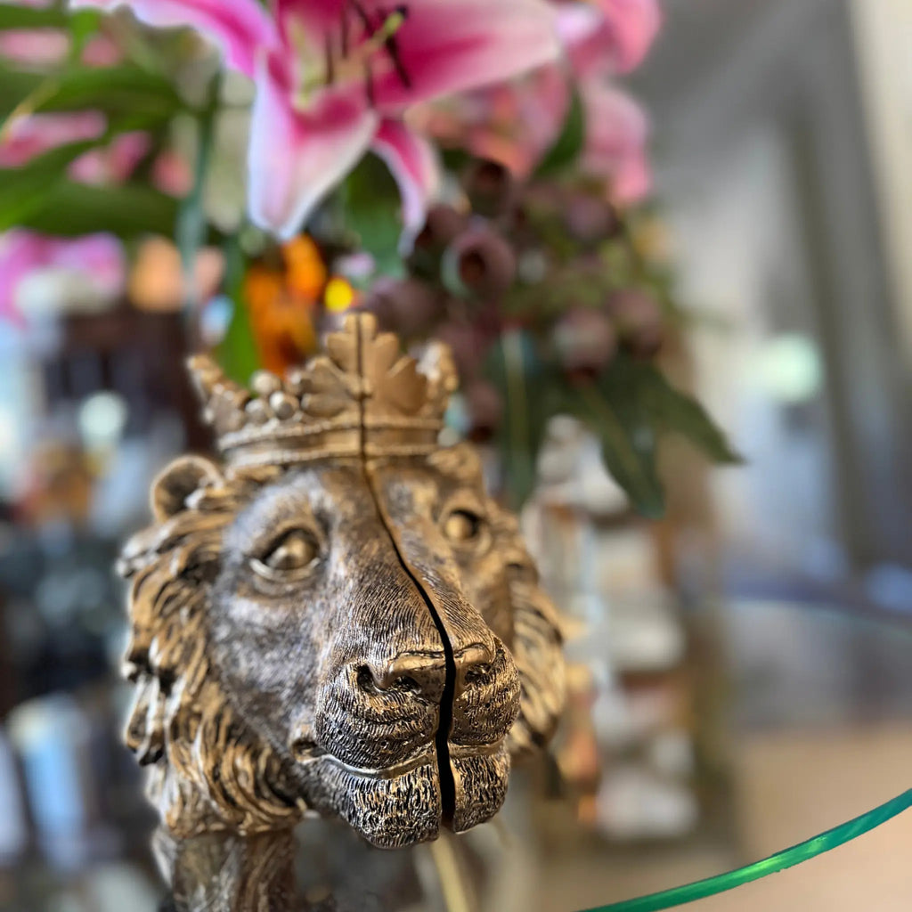 A close-up of a decorative lion head sculpture with a crown, shown in a real indoor setting with blurred pink flowers and greenery in the background.