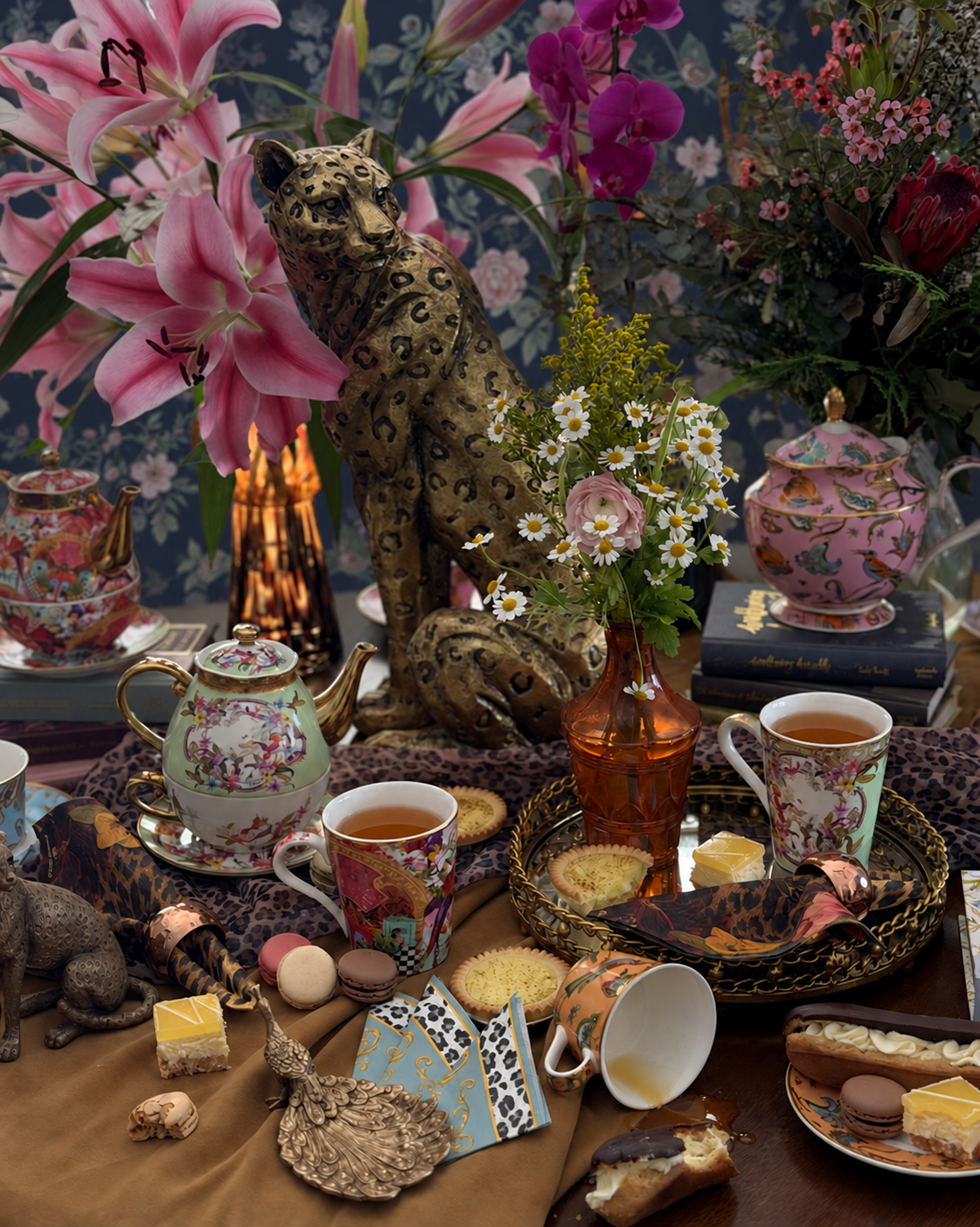 An ornate tea-time still life with floral teapots, matching teacups, pastries, flowers, and a leopard-print decorative object on a heavily styled table against a floral backdrop.