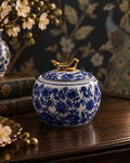 A round ceramic jar with a blue and white floral pattern and a small gold bird-shaped lid topper, displayed on a dark wooden surface with decorative books and flowers in the background.