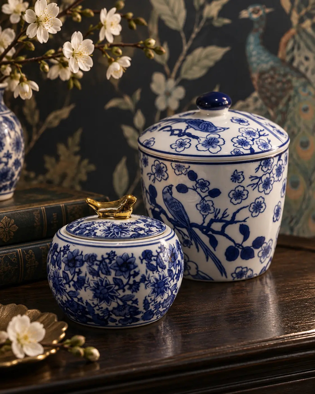 Two blue and white porcelain lidded jars with gold-toned decorative lids, displayed on a dark wooden table against a dark floral wallpaper background.